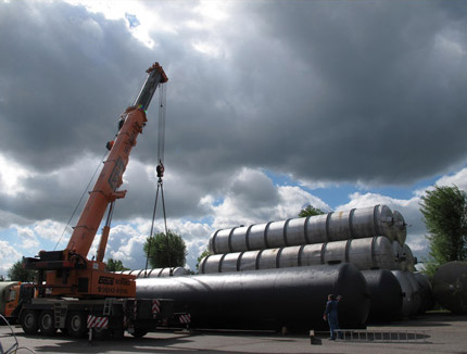 Kran hat liegenden, schwarzen Wassertank mit 170.000 Liter Volumen auf dem Au&szlig;enlager der Firma Tank und Apparate Barth GmbH an den Kran&ouml;sen eingeh&auml;ngt; daneben liegen &uuml;bereinander gestapelt VA-Beh&auml;lter mit 103.000 Liter Volumen